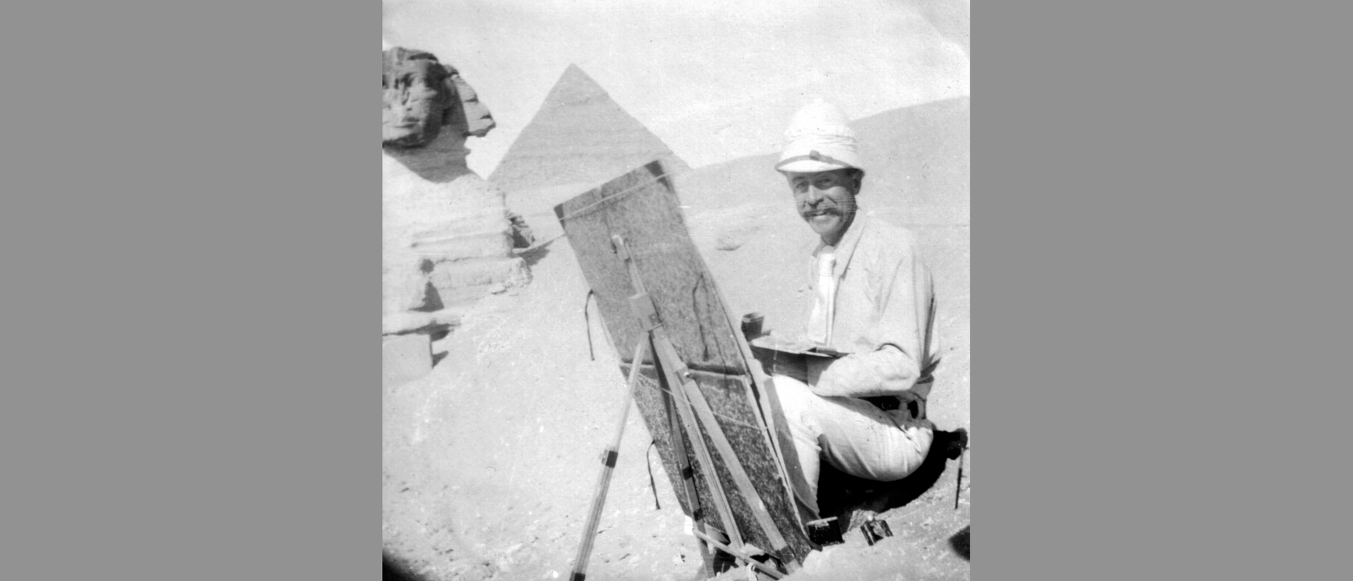 smiling man wearing pith helmet, holding palette in front of easel with sphinx and pyramid in background.