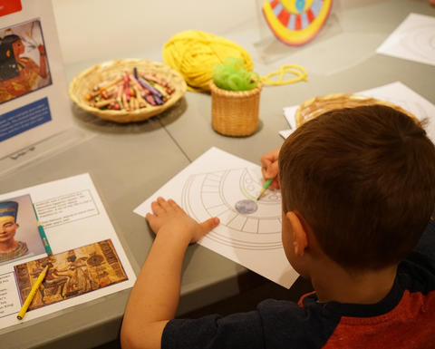 A child colors in an outline of an Egyptian collar while looking at images of Egyptian art.